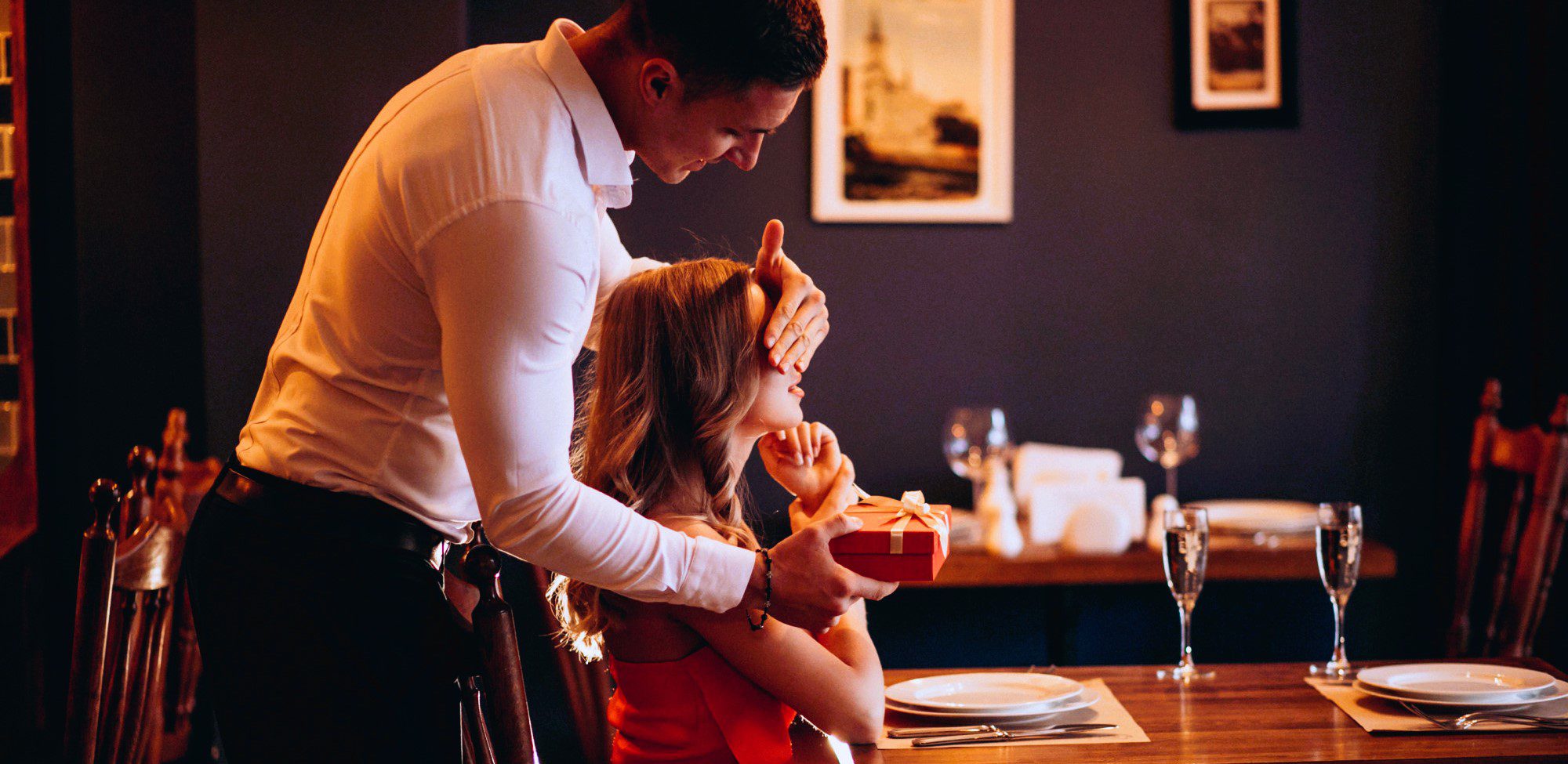 elegant couple elegant couple in restaurant, her sitting at the table, him standing behind her, holding her eyes closed and offering her a present as surprise